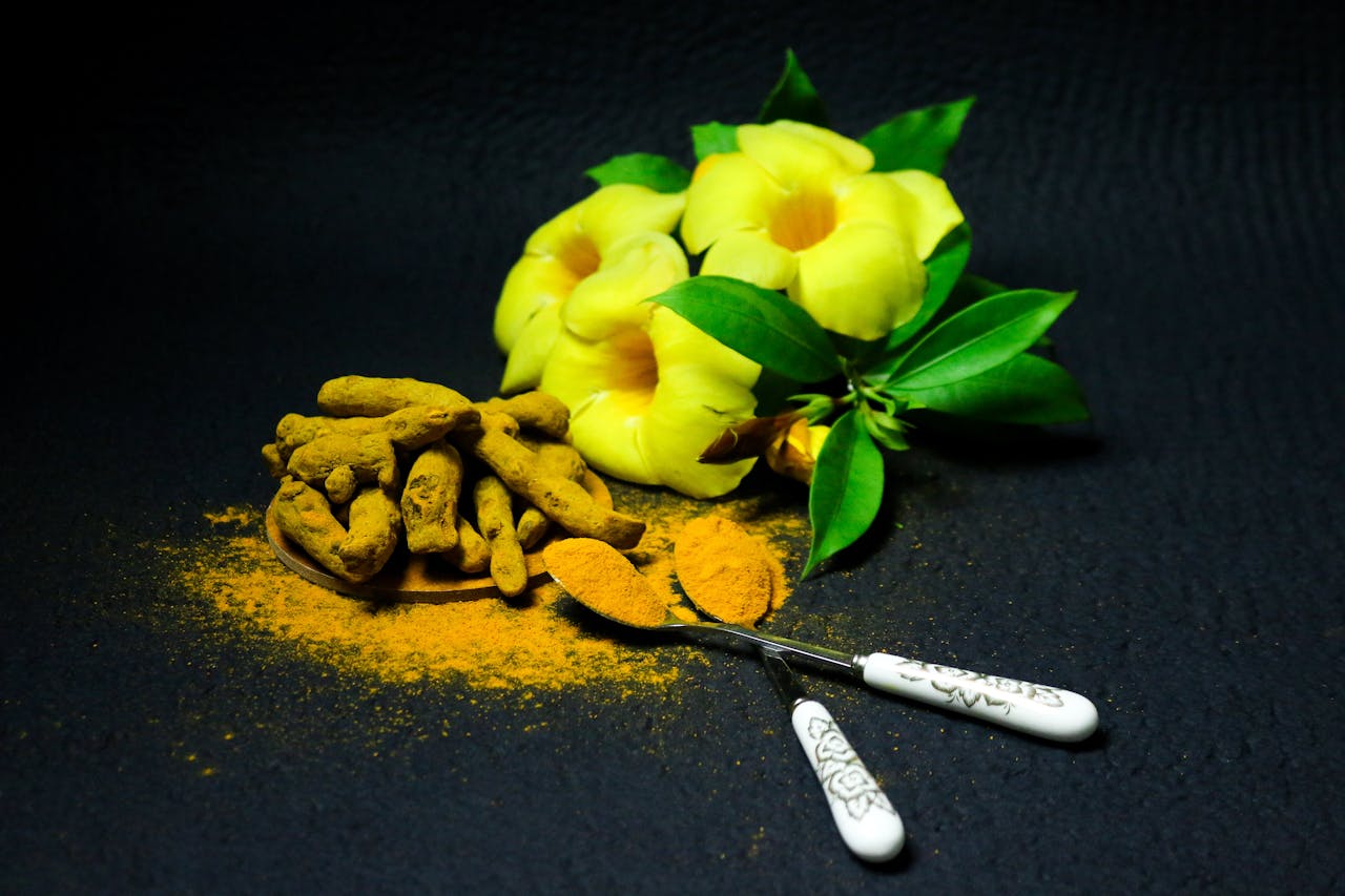 Vibrant turmeric powder and roots with yellow flowers on a dark background, showcasing spices and natural elements.